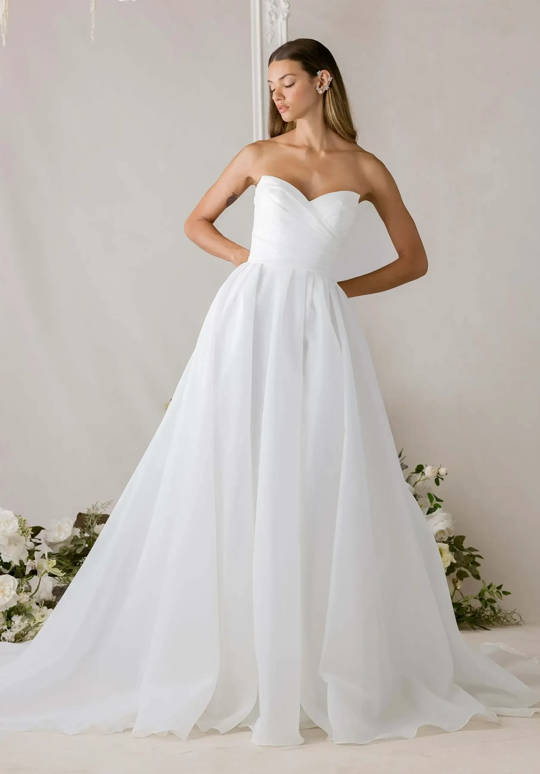 A model wearing a strapless white gown, standing gracefully with one hand on her hip, against a neutral background decorated with flowers.