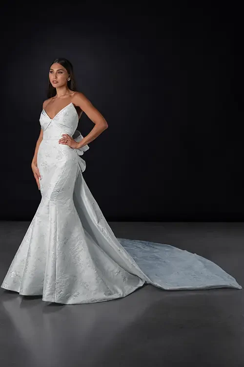 A model wearing a sleek, fitted light blue gown with a mermaid silhouette and a long train, posed against a dark background.