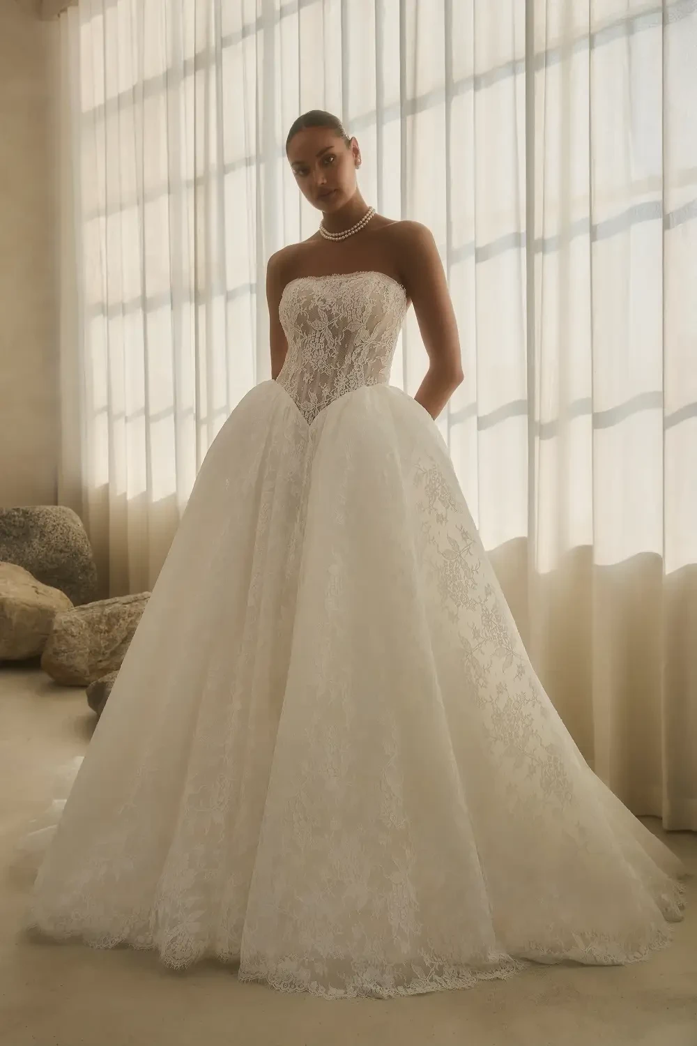 A model standing in a strapless, intricate white wedding gown with a voluminous skirt, in a softly lit room with sheer curtains.