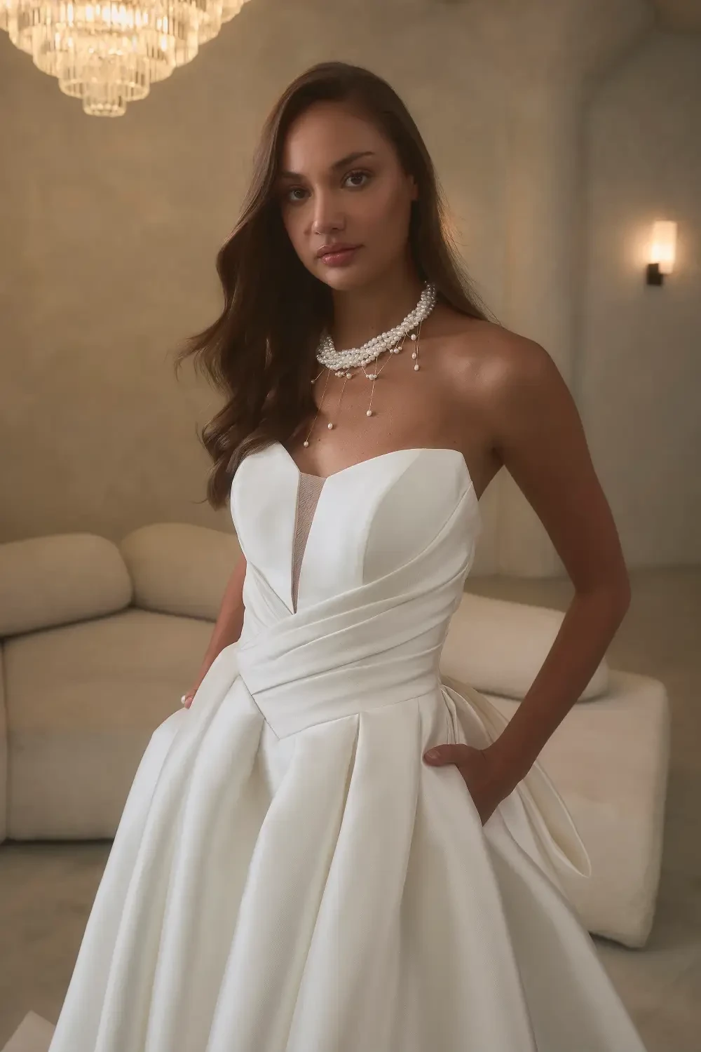 A model wearing a white strapless gown with a detailed bodice and flowing skirt, standing in a softly lit room.