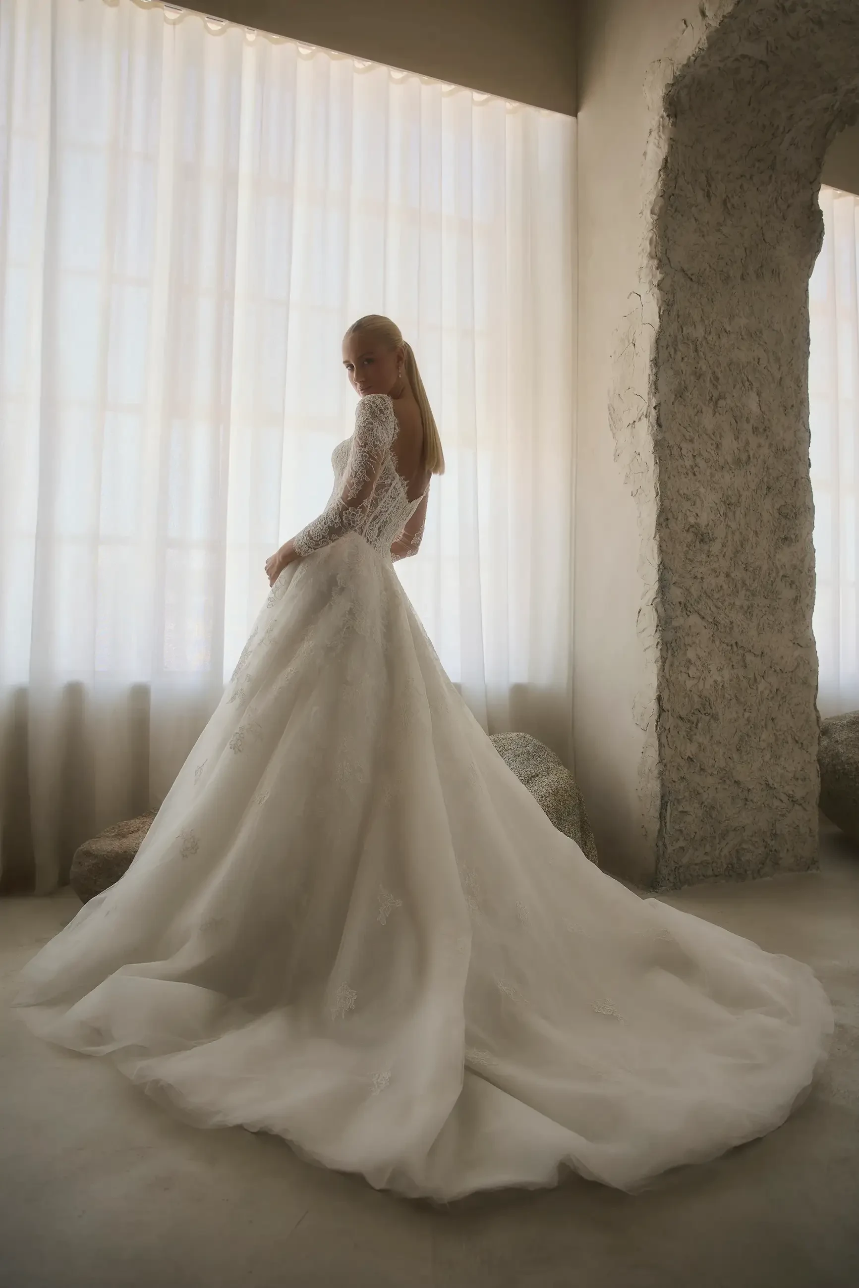A woman in a long, flowing white dress stands by a window with sheer curtains, her back to the camera. The dress has long sleeves and a voluminous train.