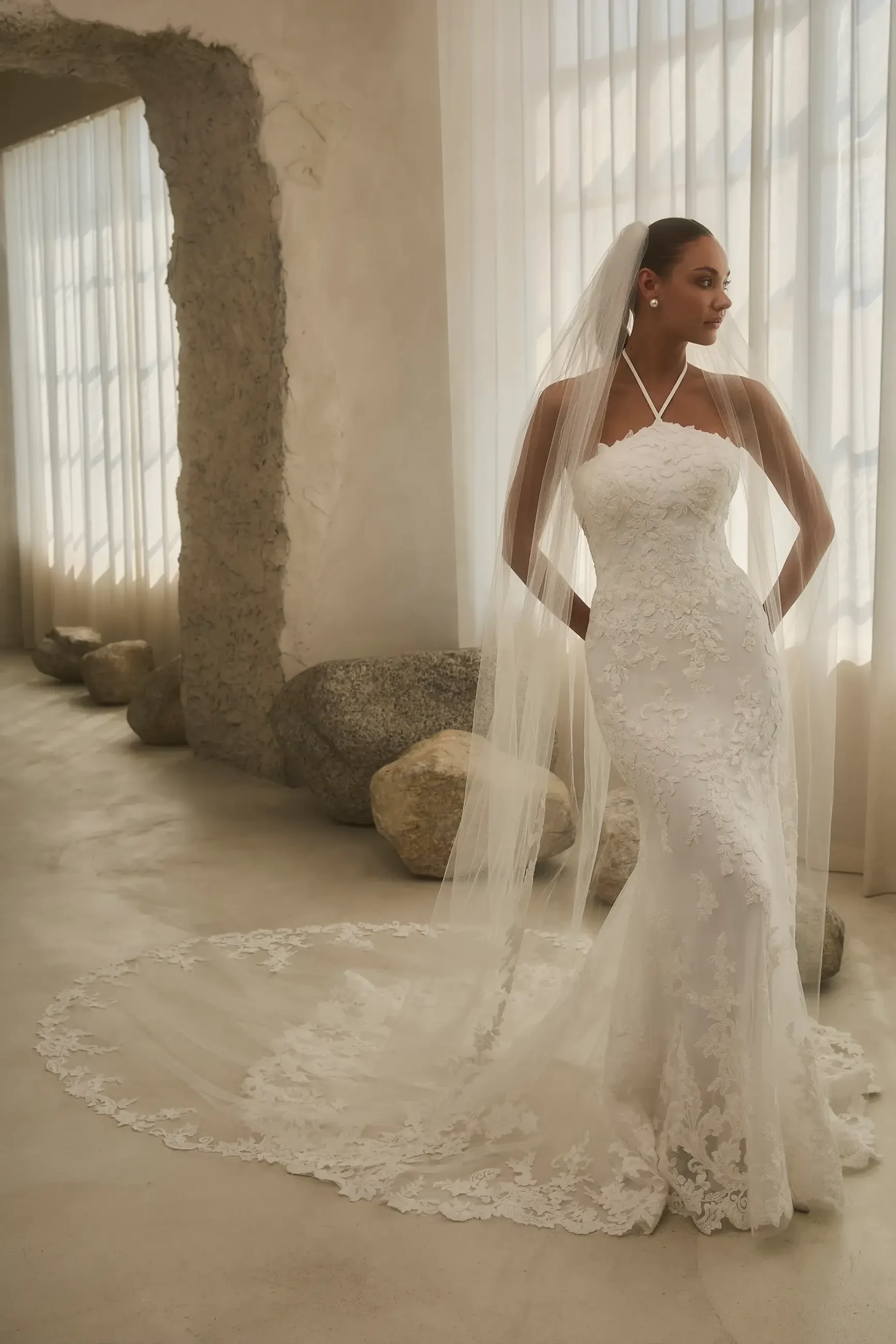A bride wearing a fitted white wedding dress with lace detailing and a long veil, standing gracefully in a bright, airy space with large windows and smooth stone decor.