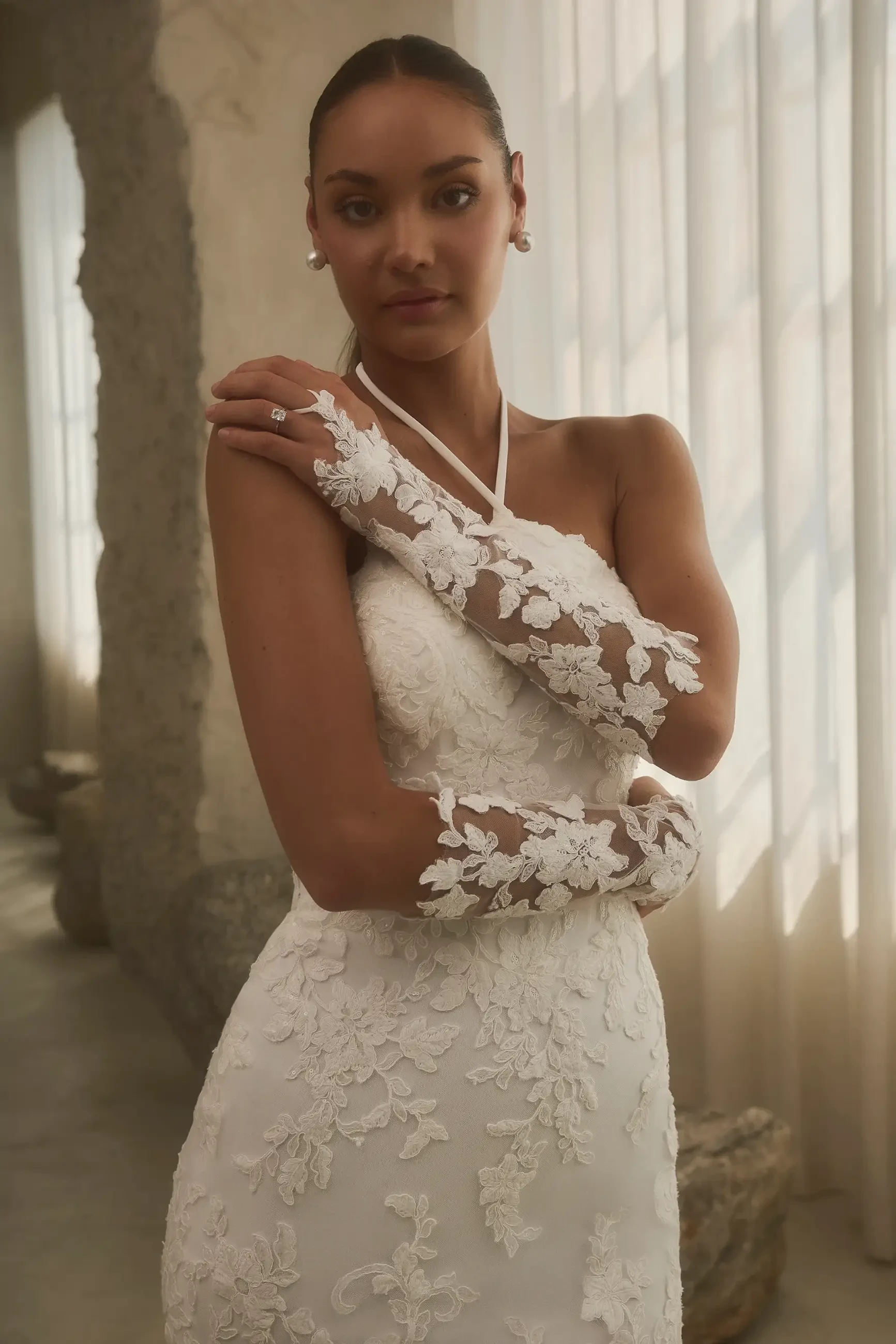 A model wearing a white wedding dress with floral embellishments and long lace gloves, standing against a softly lit background.