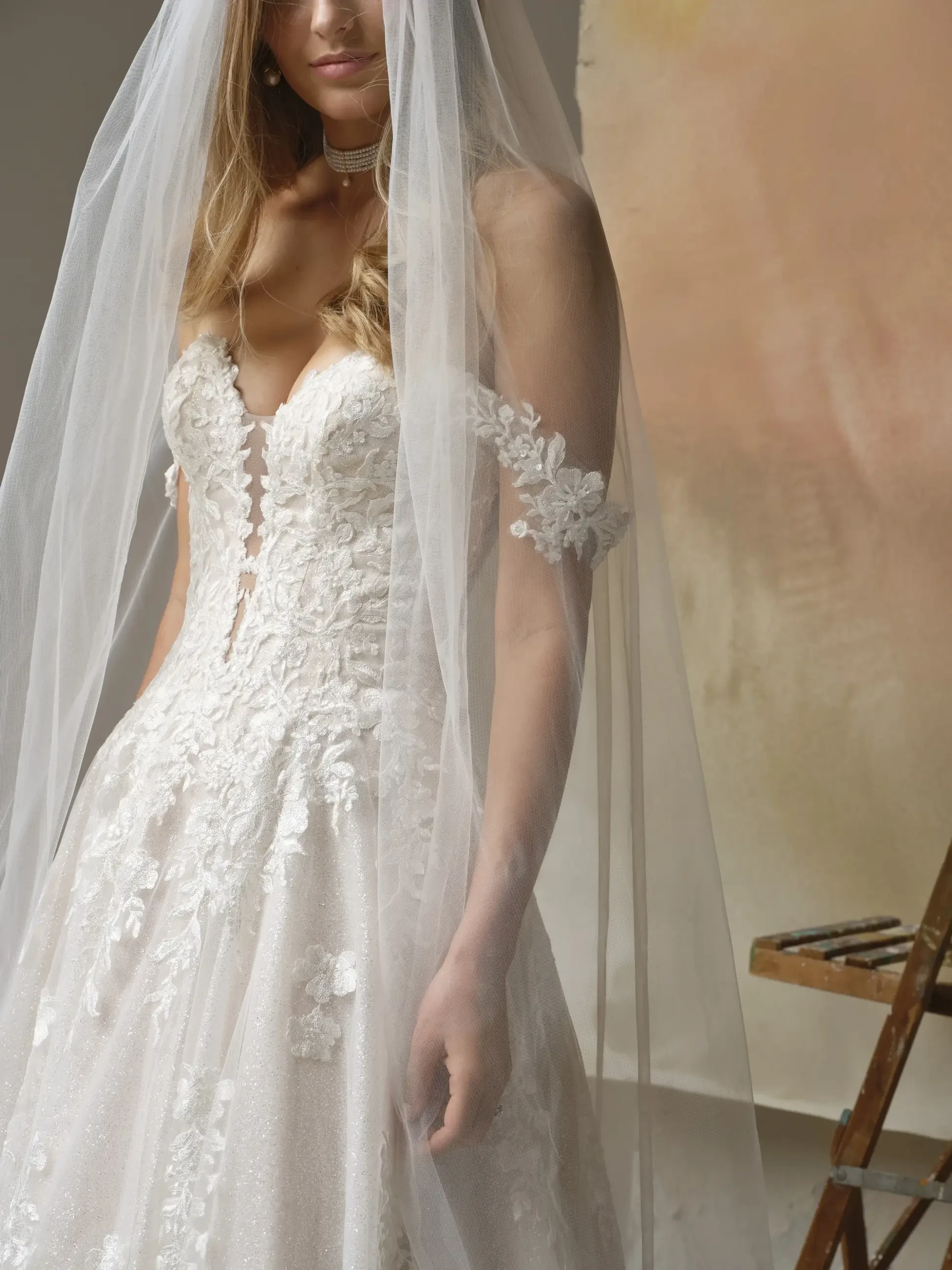 A close-up of a wedding dress with intricate floral patterns and lace details, worn by a woman with long hair partially covered by a sheer veil.
