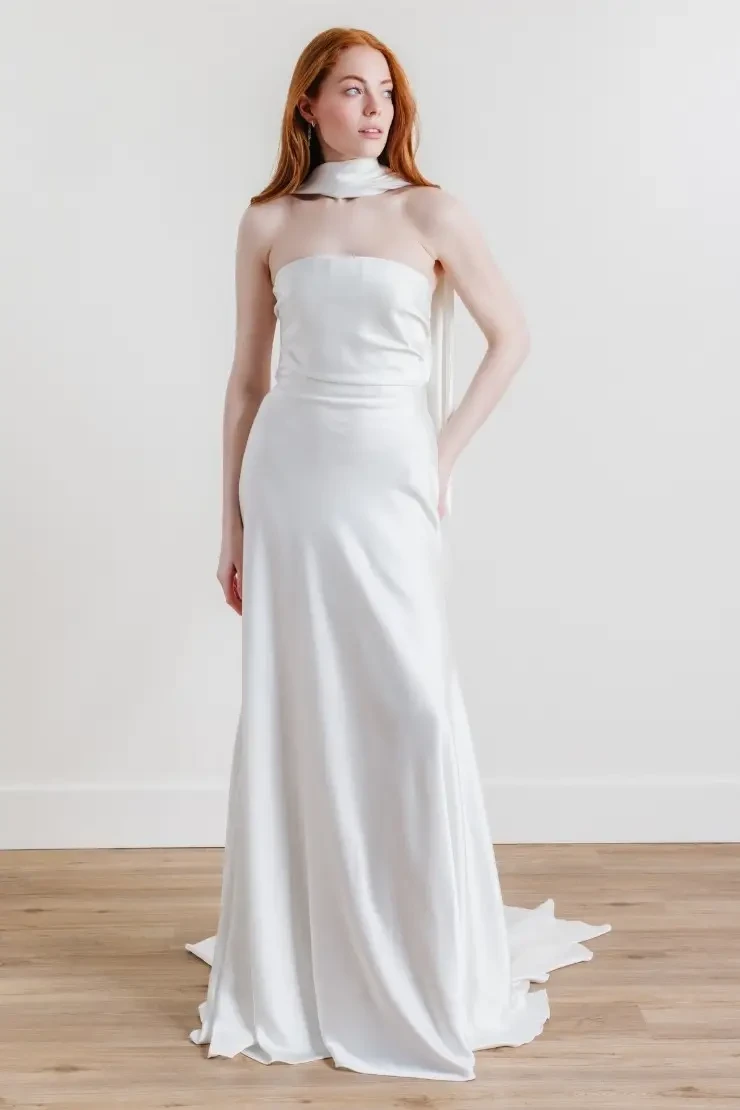 A woman in a strapless, white satin gown stands on a wooden floor. The gown has a fitted silhouette and features a long train. She has long, wavy red hair and is posing with one hand on her hip.