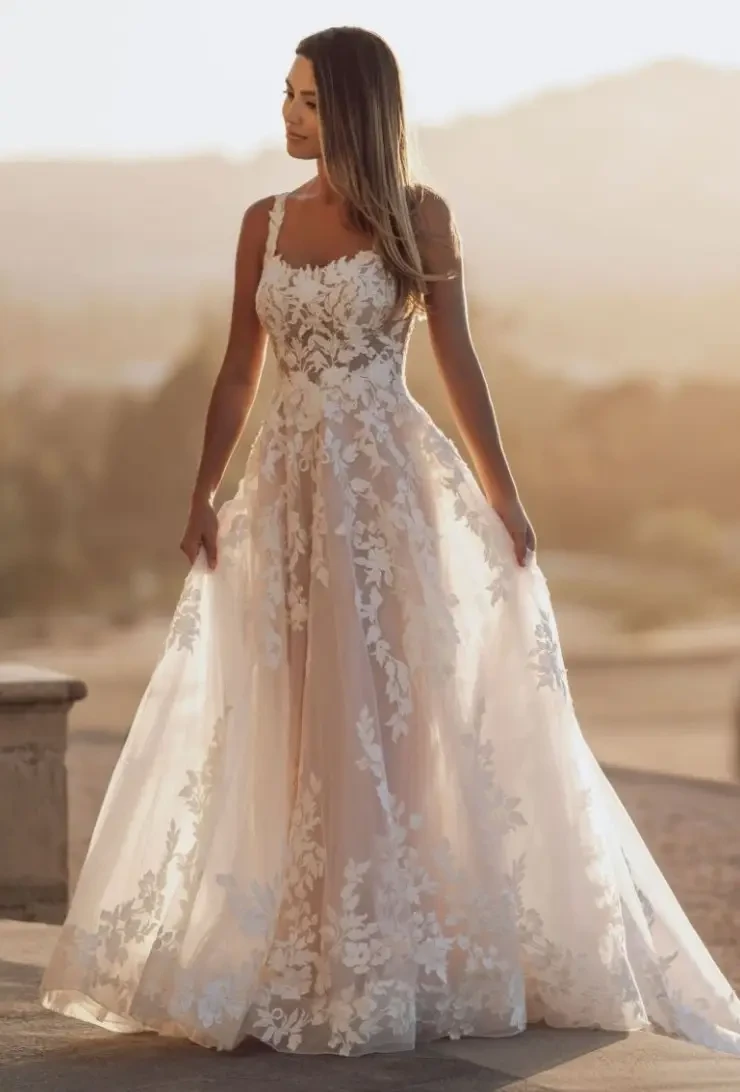 A woman wearing an elegant, floral embroidered gown stands outdoors, illuminated by sunlight with a soft landscape in the background.