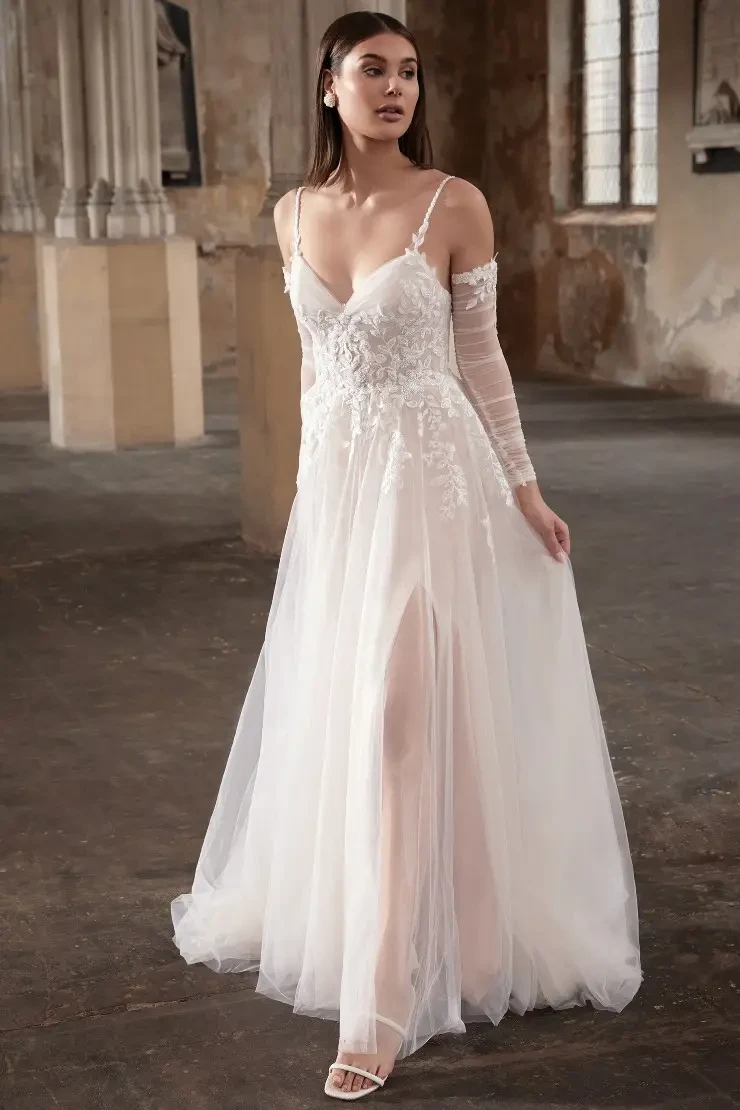 A model wearing a delicate white wedding dress with floral embellishments, featuring a deep V-neckline and a thigh-high slit, stands in an empty, rustic room with stone walls and large windows.