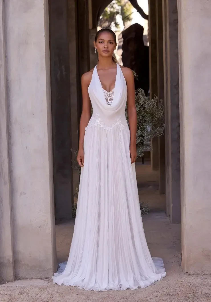 A model is wearing a long, flowing white dress with a halter neckline and intricate detailing, standing in an outdoor setting with stone structures in the background.
