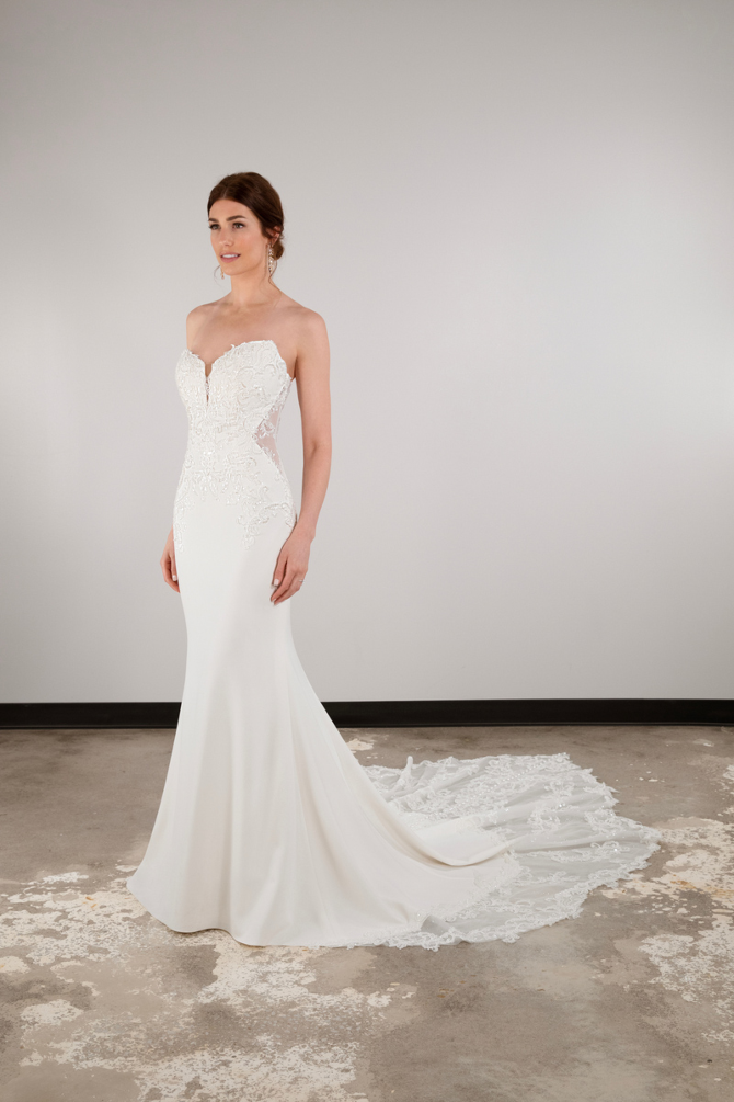 A woman in a fitted white wedding gown with lace details and a train, standing against a plain background.