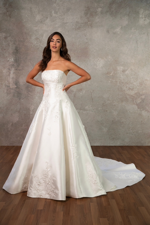A woman wearing a strapless white wedding dress with an A-line silhouette, standing gracefully with her hands on her hips against a textured gray background.