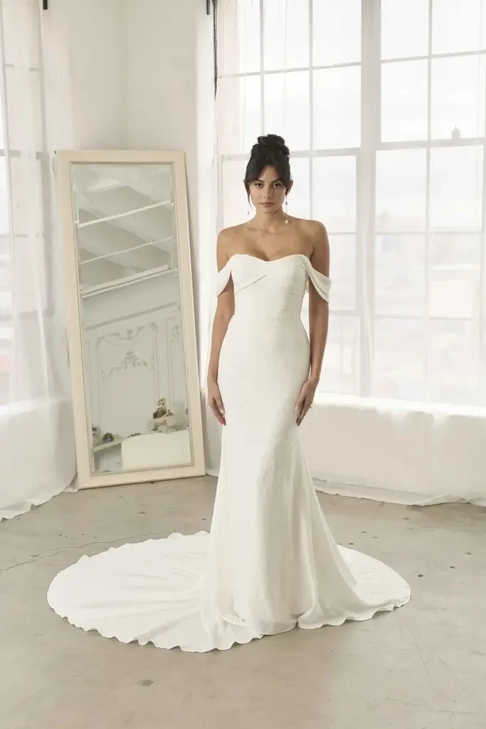 A woman in a white off-shoulder wedding dress stands in a bright room with large windows.