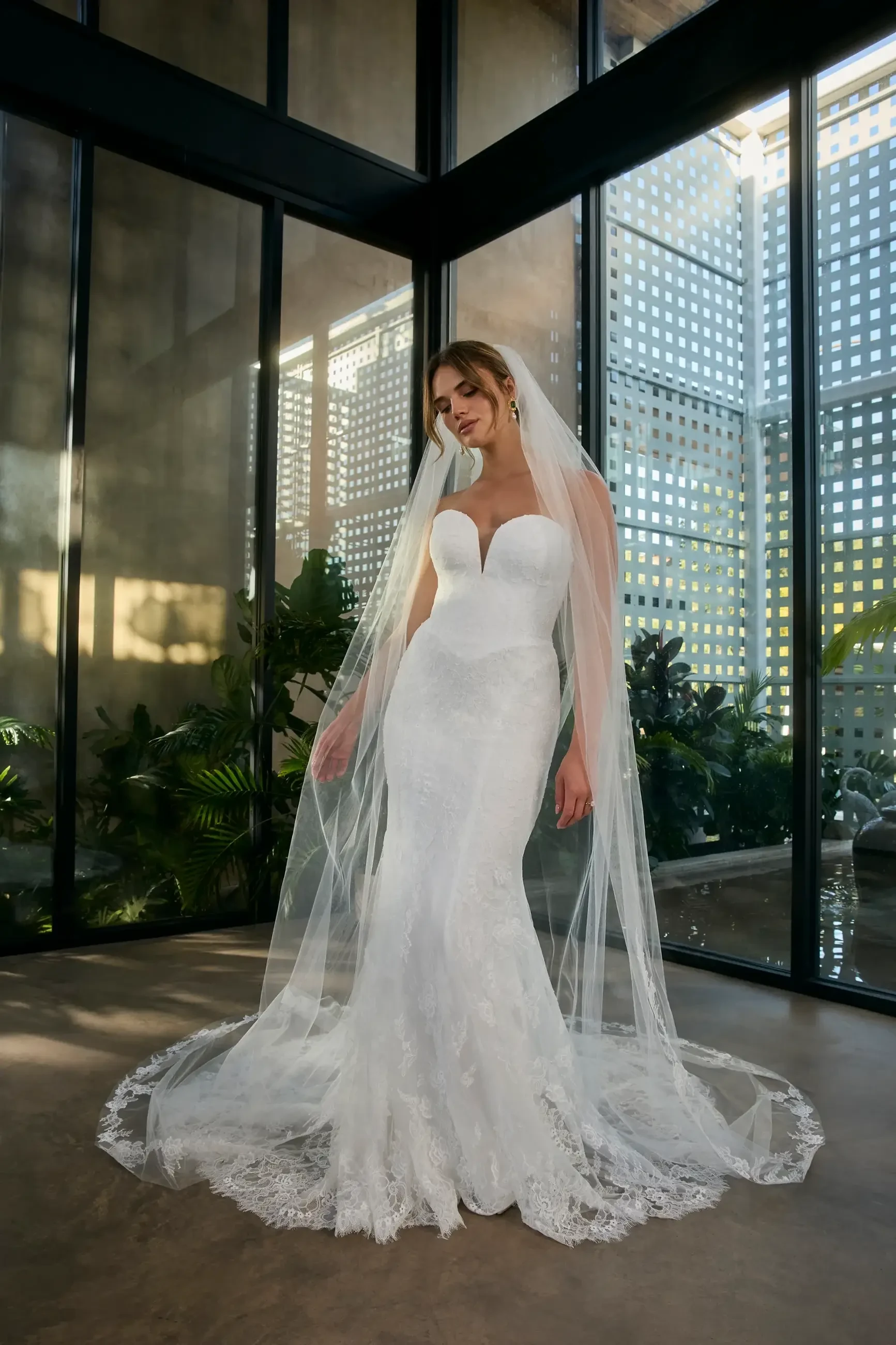 A woman in a fitted white wedding dress with a sweetheart neckline and a long veil, standing in an indoor space with large windows and greenery around her.