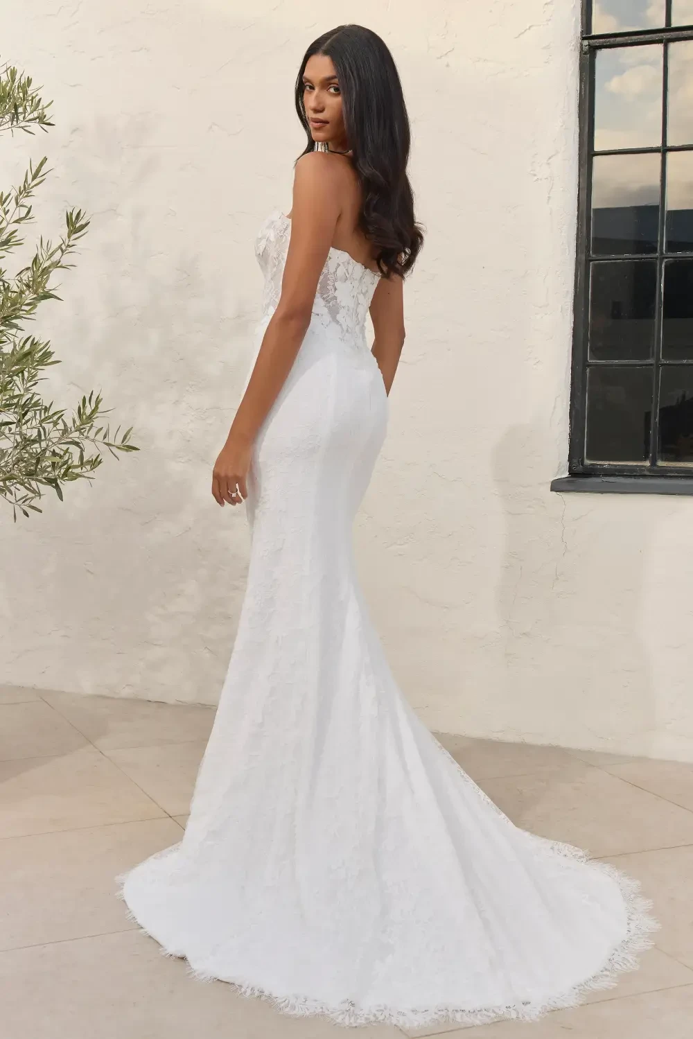 A model is wearing a form-fitting white lace wedding gown, showcasing a mermaid silhouette with a train, standing in profile against a light-colored wall and window.