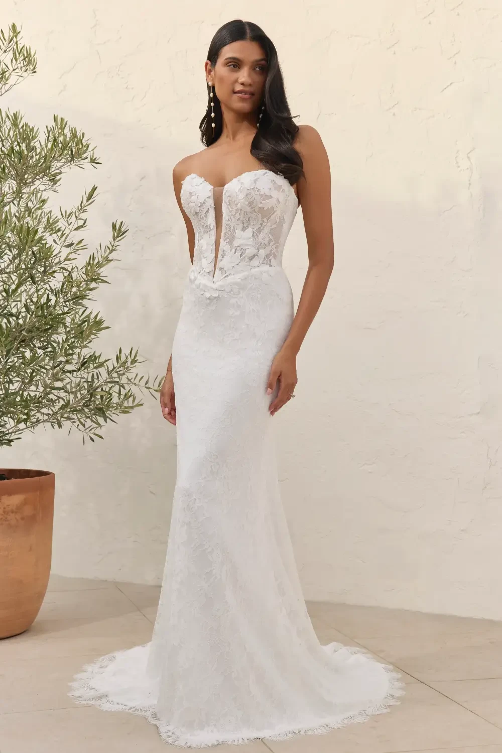 A woman in a white strapless wedding dress with a fitted silhouette and lace details, standing near a potted plant.