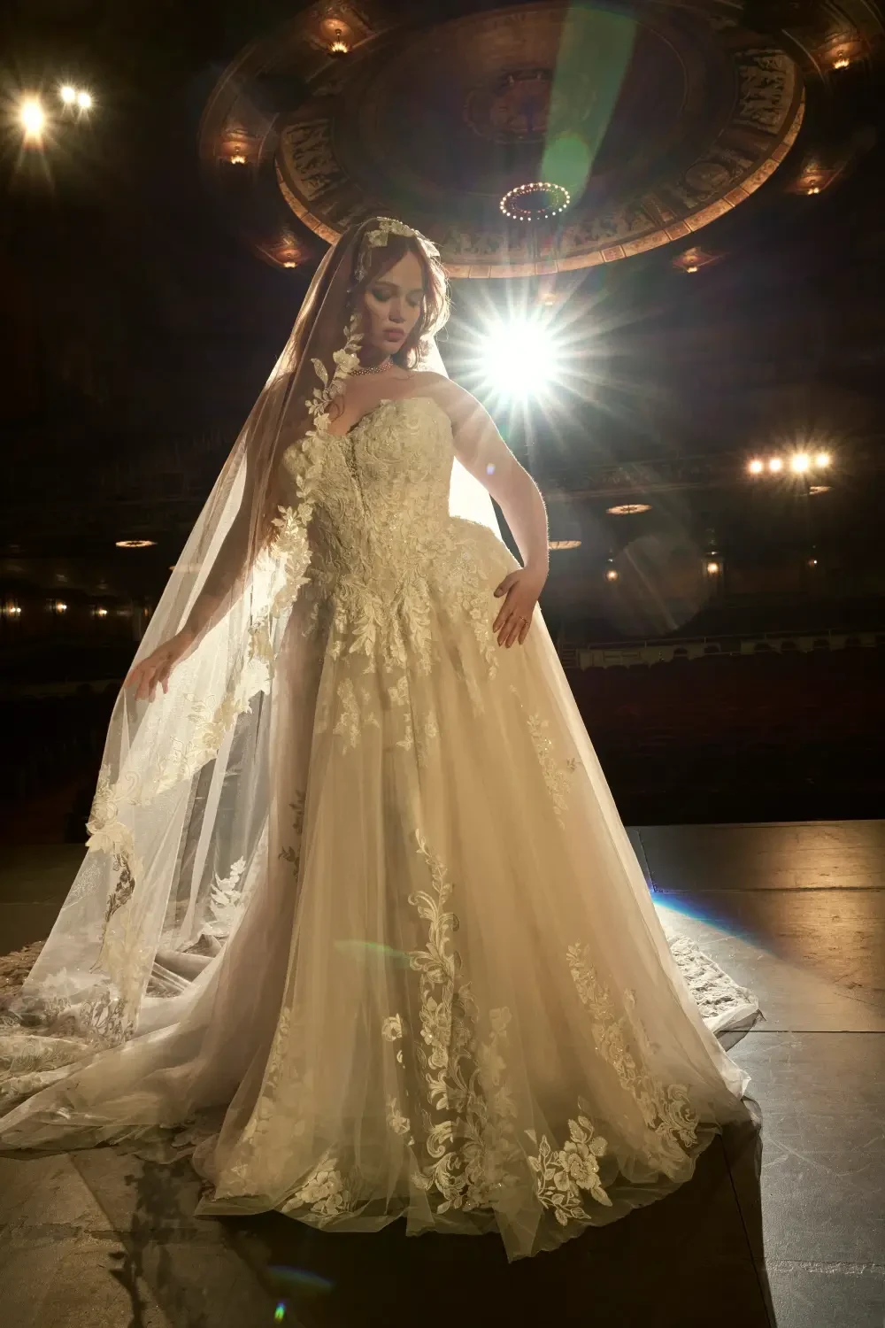 A model wearing an elegant wedding dress with intricate lace details and a long veil, posing on a stage with dramatic lighting in the background.