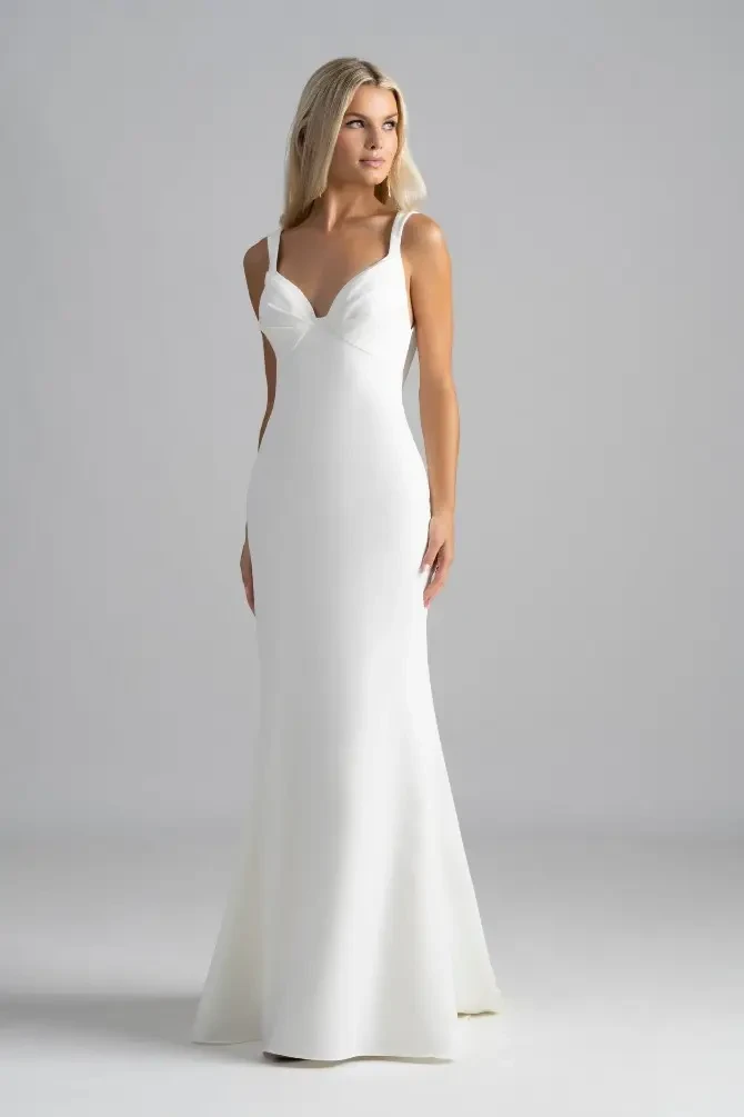 A woman wearing a elegant white dress, standing against a neutral grey background. The dress features thin straps and a fitted silhouette that flares out at the bottom.