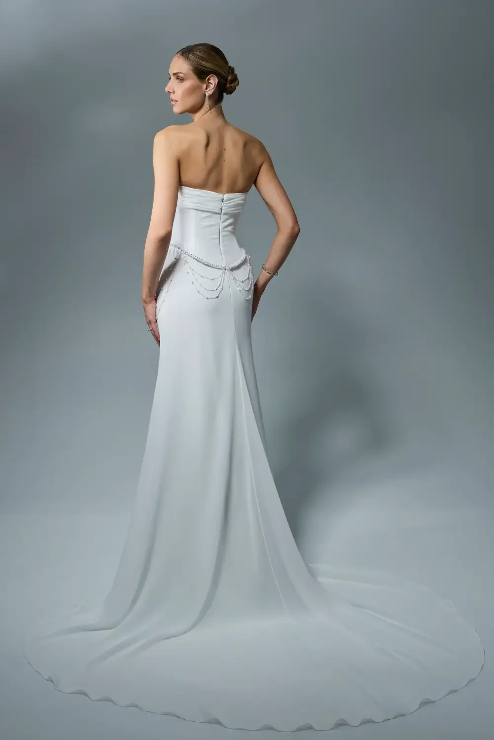 A woman in a strapless white gown with a flowing silhouette and decorative chains at the waist, standing against a soft gray background.