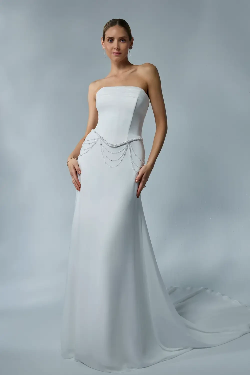 A woman in a strapless white gown with a flowing silhouette and decorative chains at the waist, standing against a soft gray background.
