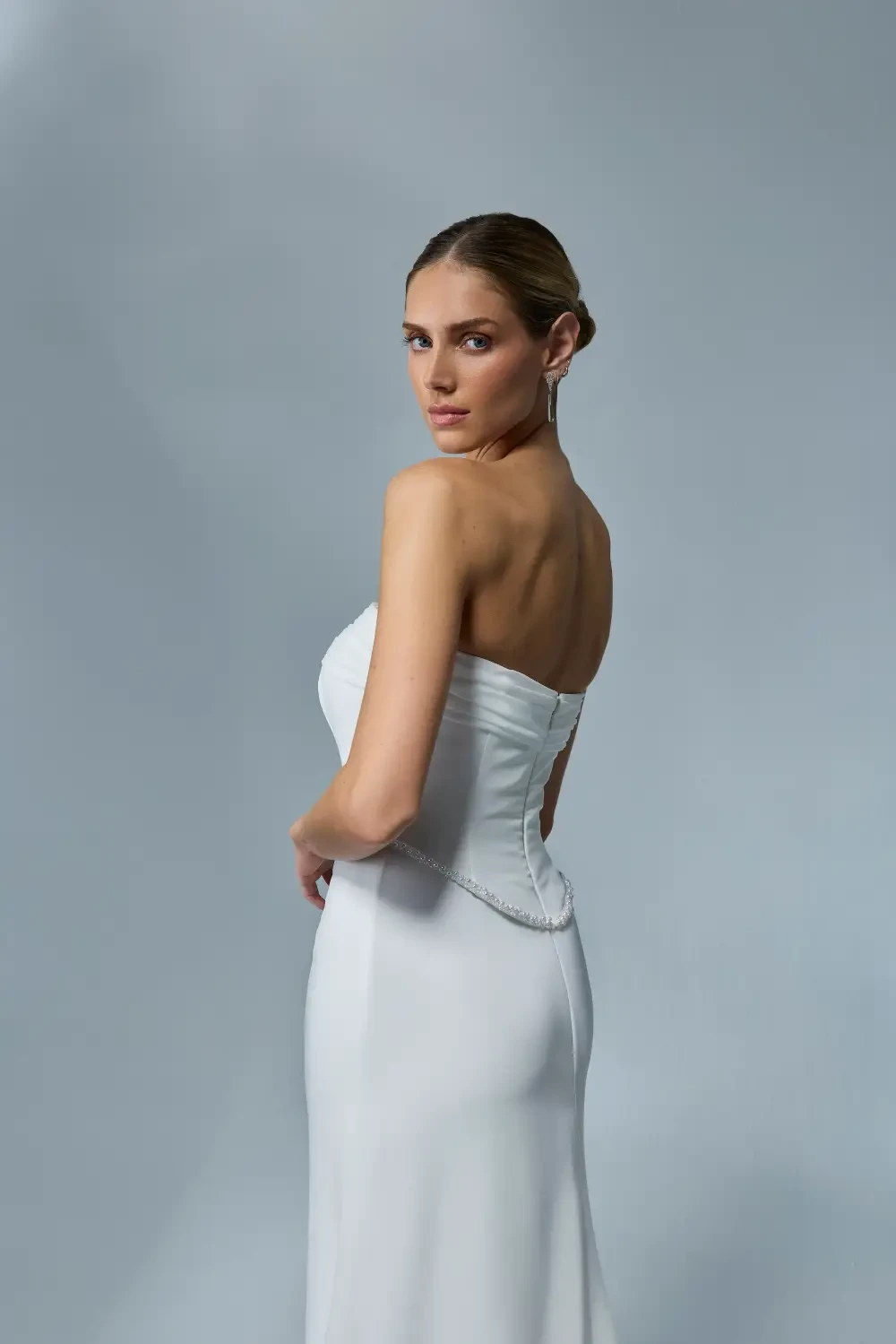 A woman in a strapless white dress stands against a light grey background, looking over her shoulder.