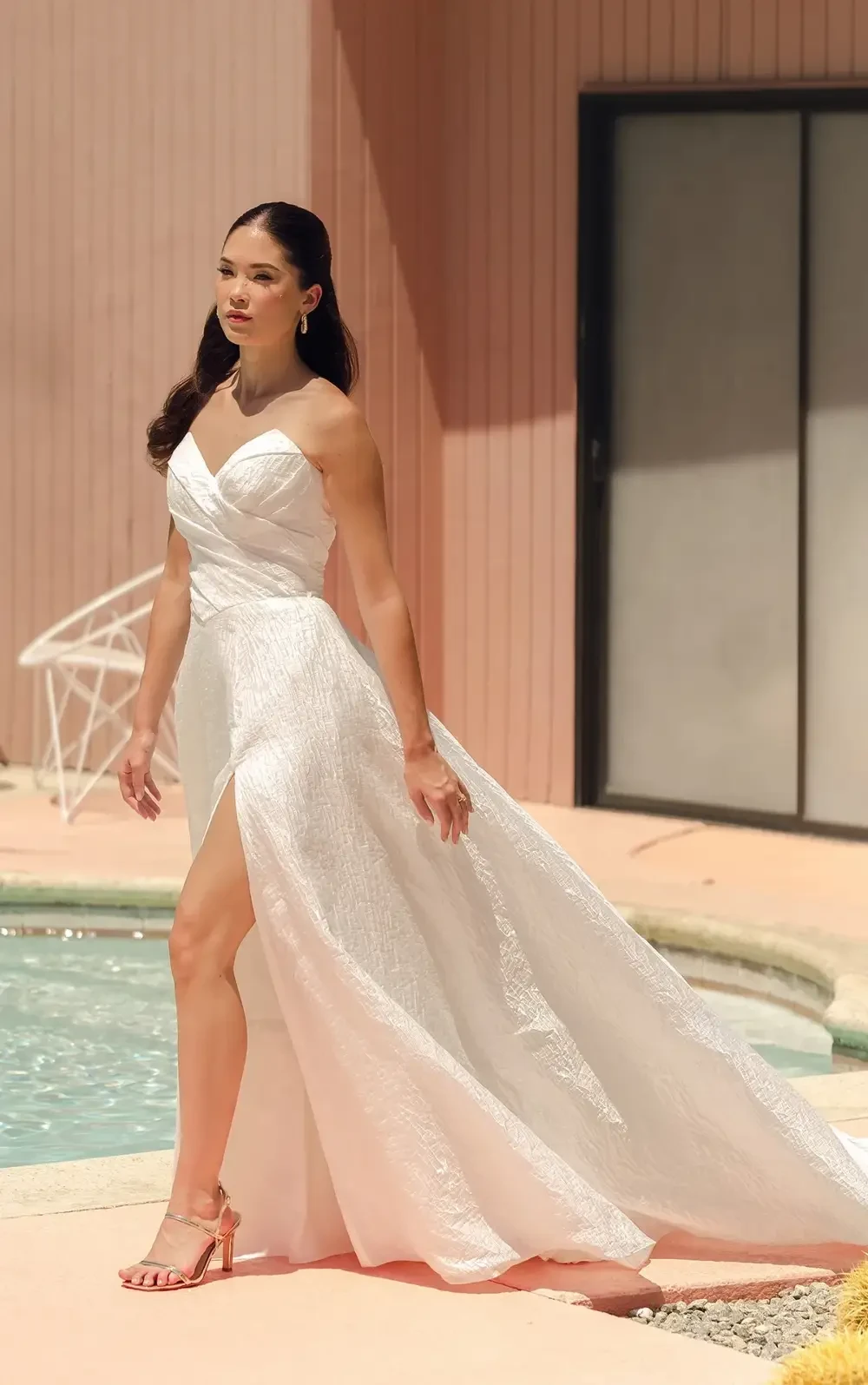 A woman in a white evening gown with a high slit stands by a pool, in front of a pink building.