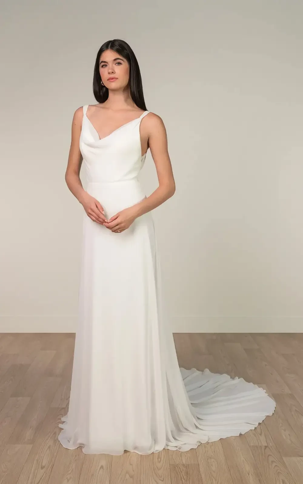 A woman stands in a white gown with a flowing train, featuring a draped neckline, against a neutral background.