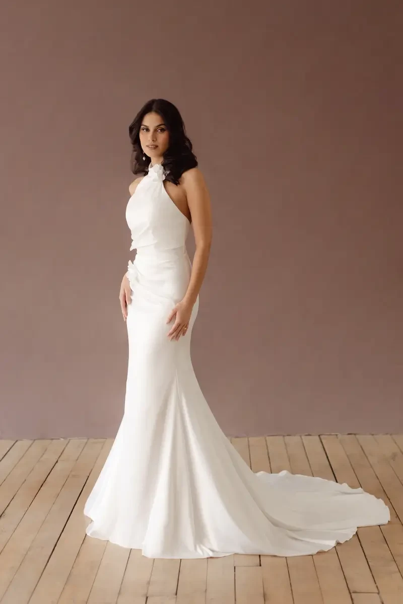 A woman in a sleek, white evening gown stands on a wooden floor against a neutral backdrop.