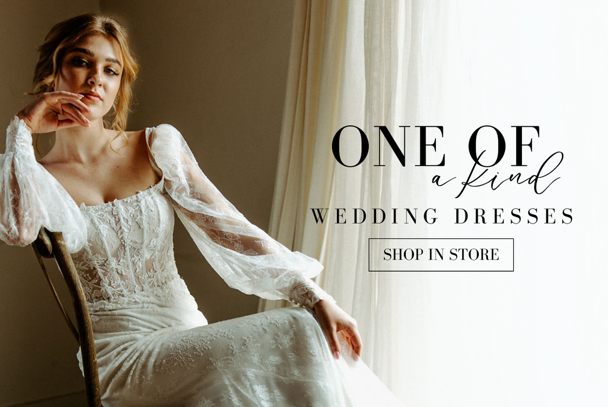 ONE OF A KIND WEDDING DRESSES SHOP IN STORE. BRIDE WEARS LONG SLEEVES LACE WEDDING DRESS WITH SHEER BODICE AND IS SEATED IN A VINTAGE CHAIR WITH WHITE CURTAINS BEHIND HER, LEANING ON HER HAND WHILE HER OTHER ARM IS OUTSTRETCHED ALONG HER CROSSED LEGS.